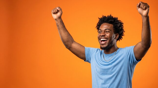 Joyful excited African American man celebrating success and triumph with raised arms against a solid bright orange background