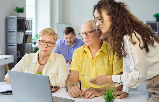 Seniors adults students with instructor training class. Instructor helps two seniors at a laptop, explaining steps and building confidence. Clear concept of adult learning and upskilling.