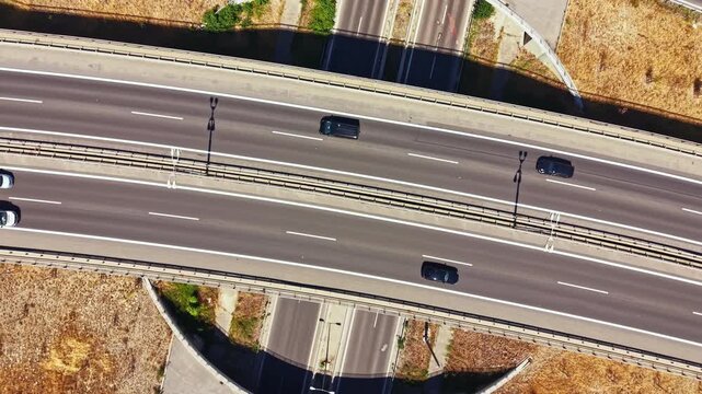 A white car drives on a highway with multiple lanes. The view shows the road layout and surrounding areas. The scene features clear weather and dry grass nearby.