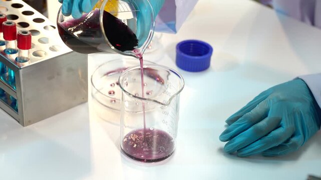 Laboratory worker in blue gloves pours purple liquid from a flask into a glass beaker beside test tubes and lab equipment.