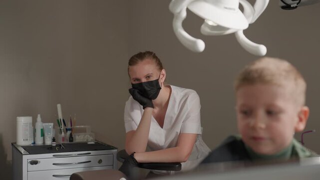 Close up of dental hygienist in white uniform and black gloves resting at her post observing out of focus young boy seated nearby. Dental tools like toothbrushes are visible on counter