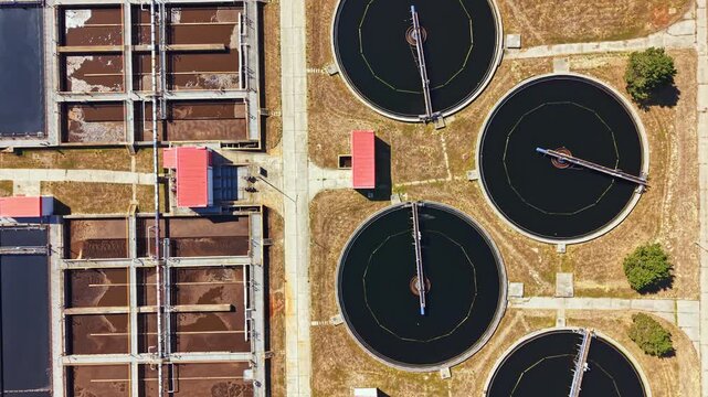 A water treatment facility is shown from above. It includes large round tanks filled with dark water and rectangular sections alongside them. The scene takes place under clear skies during the day.