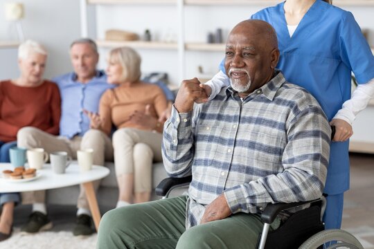 Happy Elderly Black Man Sitting In Wheelchair, Smiling And Engaging With Group Of Three In A Comfortable Healthcare Facility, Promoting Community Interaction And Support In Senior Care.