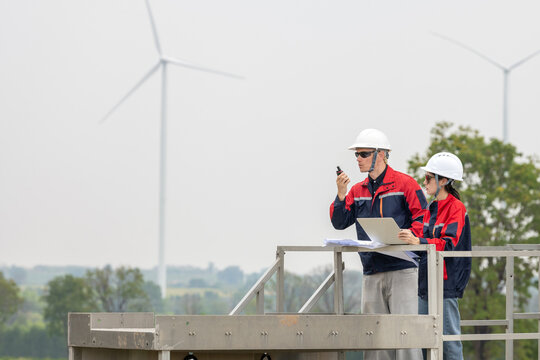 Two renewable energy engineers in safety gear discussing with laptop radio at wind turbine field. Male and female inspection engineer team working at windmill site for sustainability project.