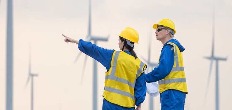 Two renewable energy engineers discussing with blueprint while pointing away at wind turbine field. Male and female inspection engineer team working at windmill site for sustainability project.