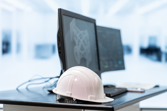 White safety helmet on a desk beside dual computer monitors in a bright modern office, representing engineering, construction planning, workplace safety and design work.