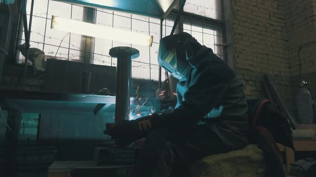 Welder Working on Industrial Pipe: Low Angle Shot of Worker in Protective Mask with Sparks Flying | Steel Water Pipeline Welding in Factory Workshop | Cinematic Industrial Scene with Brick Wall