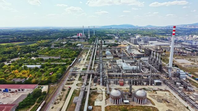 A large industrial site is seen from above. There are many pipes, tanks, and structures. The facility is surrounded by greenery and hills in the distance. Clouds float overhead.