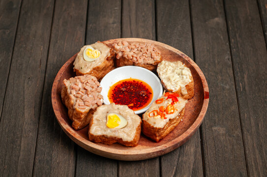 a wooden plate of tahu bakso (tofu stuffed with meat paste) served with various toppings and a dipping sauce.
