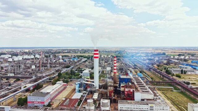 An industrial area with tall smokestacks releasing smoke into the air. Various buildings are spread across the flat land. The sky is mostly cloudy with patches of blue.
