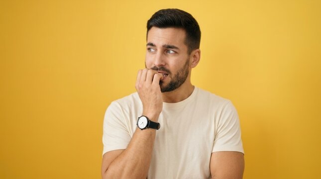 Hispanic man looking nervous and stressed, biting nails with hand on mouth over yellow background.