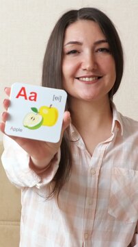 Young female teacher holding up a flashcard with the letter a and a picture of an apple, smiling at the camera during a remote english language lesson for preschool children