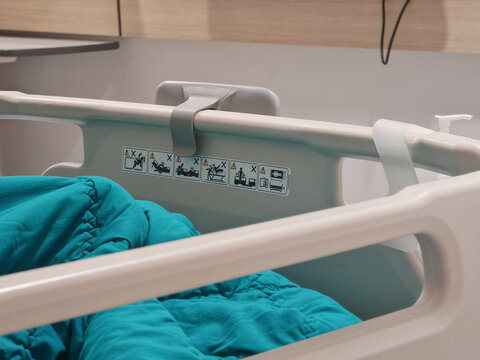 High-resolution close-up photograph of a modern ergonomic hospital bed side rail featuring clear safety warning pictograms and a teal medical blanket. Ideal target for healthcare facility brochures