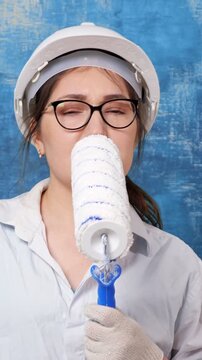 Playful female painter wearing a protective hard hat and glasses singing with passion, using a clean paint roller as a microphone for a creative and fun performance during her break