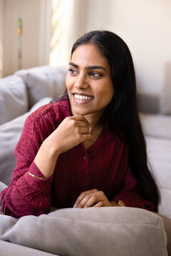 Young dreamy Indian woman sitting on couch, smiling, gazing aside, looks calm, thoughtful, and contented, enjoying quiet moment, pleasant relaxation alone at home. Happiness, peaceful domestic life