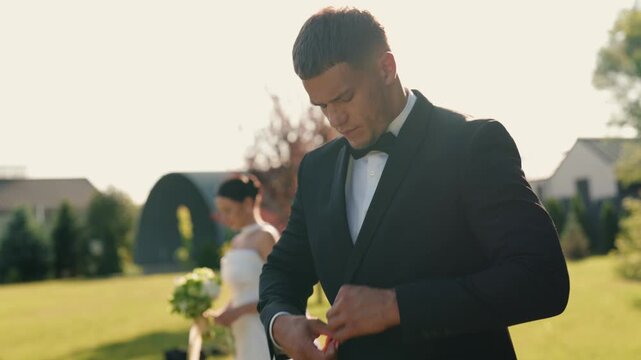 Handsome groom in a black tuxedo getting ready for the wedding ceremony while his bride waits in the background on a sunny day. This romantic slow-motion video captures a beautiful moment