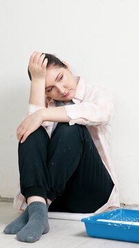 Upset female painter feeling overwhelmed and tired during apartment repair and renovation, sitting on the floor leaning against a freshly painted wall with a blue tray nearby