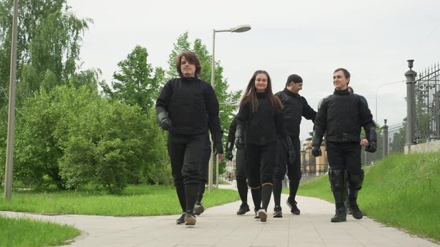 Group of people in black padded clothing walking along paved path with green grass and trees wearing protective gear including gloves and knee pads. Scene features outdoor park environment