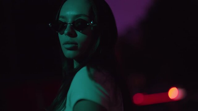 Beautiful young woman wearing fashionable sunglasses turning her head to look at the camera in slow motion, with cinematic neon lights creating a mysterious and cool urban vibe at night