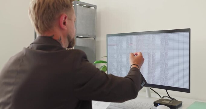 Businessman analyst accountant in office reviewing spreadsheet data. He points at the monitor, explaining figures for finance analysis, audit, and taxes. Clear financial data analysis concept.