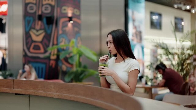 Beautiful young woman sitting at a bar counter inside a shopping mall and drinking a healthy green smoothie with mint leaves through a straw, enjoying her refreshing detox beverage