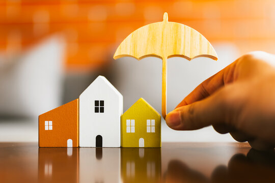 Property insurance and real estate protection concept with hand holding wooden umbrella over miniature house models on table with warm brick background for mortgage safety and coverage.