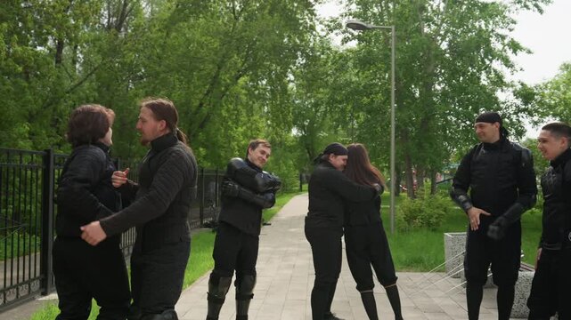 Close up of multiple people in black protective gear practicing physical restraint techniques outdoors on paved path surrounded by green trees. Some people are restrained while others