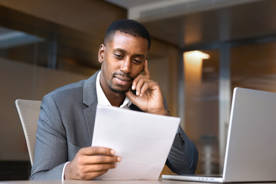 Focused Black businessman reviewing business documents, learning contract terms, studying corporate paperwork information, reading project documentation sitting at office workplace desk with laptop