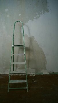 Lonely green stepladder standing inside a messy, abandoned room during apartment renovation or reconstruction, with a shadow moving across the unpainted and dilapidated concrete wall. Vertical shot.
