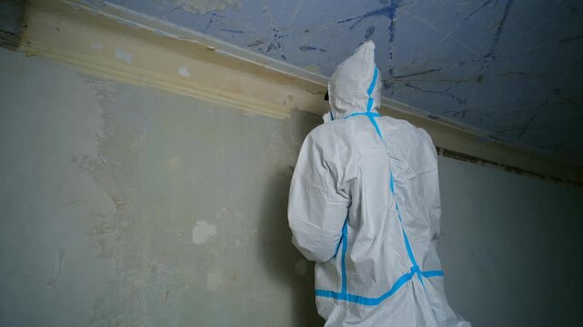 Professional plasterer wearing a white protective suit and hood carefully applying putty with a trowel, leveling the surface of a wall during a room renovation and preparing it for painting. 