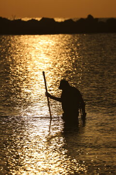Detectorist probes the seabed with a rod in golden dawn water, tools in silhouette