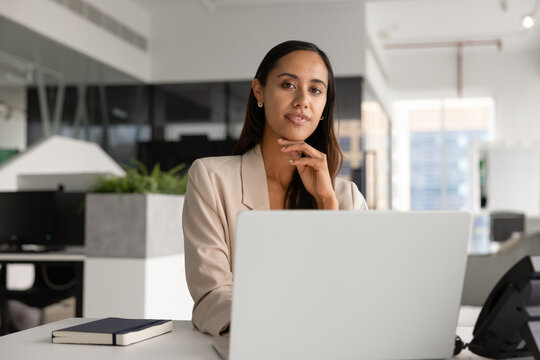 Office portrait of adult 30s businesswoman sits at workplace desk with laptop, posing, looking at camera, representing executive leadership and corporate professionalism, technology use in business
