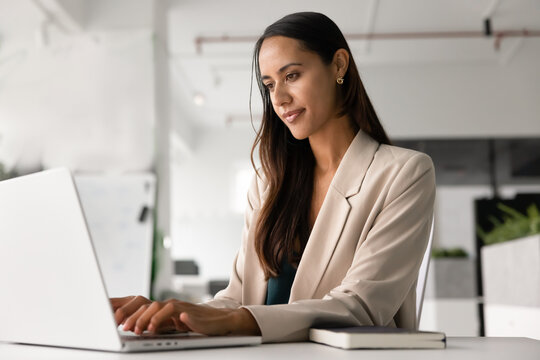 Professional woman sits at workplace desk, focused on working on laptop in corporate office, typing on computer, responding to emails. Tasks management, online workflow, technology use in business