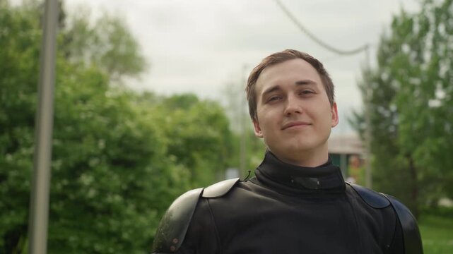 Close up of man in black protective armor standing outdoors with trees and greenery. Man adjusts his headgear revealing interior padding while maintaining steady posture throughout scene