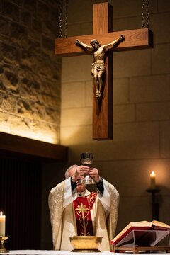 Priest holds holy chalice during mass with crucifix overhead. Man in vestments blesses communion cup before altar. Lit candle and open book complete scene.