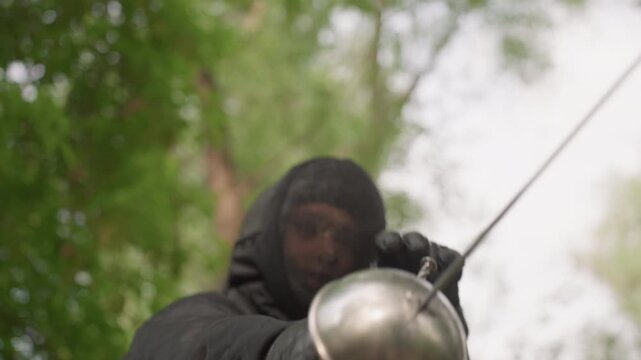 Close up of person in black fencing gear holding and adjusting their sword outdoors with trees visible. person is seen preparing for or practicing fencing focusing on weapon against natural