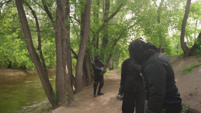 Close up of multiple people in black fencing gear preparing for match along tree lined path near water. group interacts with each other holding swords and wearing protective helmets