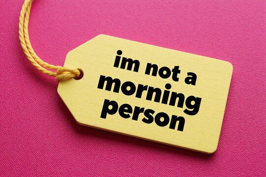 Yellow tag with black text im not a morning person rests on textured pink surface. Tag hangs by yellow string. Humorous statement relates to sleep and waking.