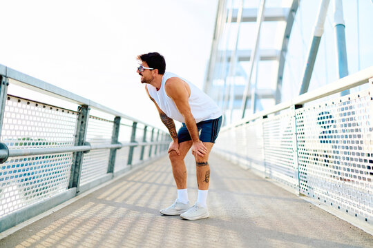 Male runner bent forward resting on modern bridge after intense urban workout, fatigued but determined, sunlight highlighting sweat and effort