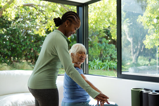 Diverse female aide in athletic top assisting Senior woman stretching at home near rolled mats