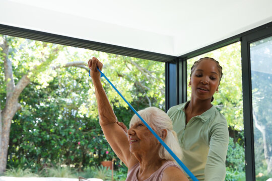 Senior woman lifting blue band African American female caregiver guiding in green top in sunroom