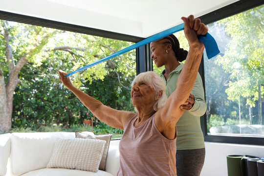 Senior female with African-American trainer stretching with blue band on white sofa in living room