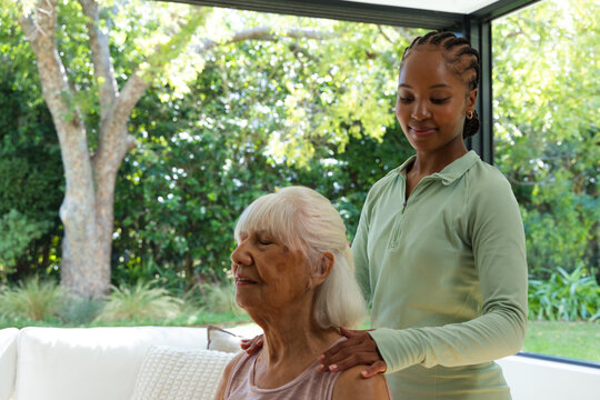 Senior woman sitting on sofa African American woman carer standing behind wearing ziptop by garden
