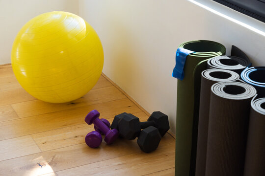 Yellow stability ball resting on wood floor left, purple dumbbells lying, mats stacking, copy space