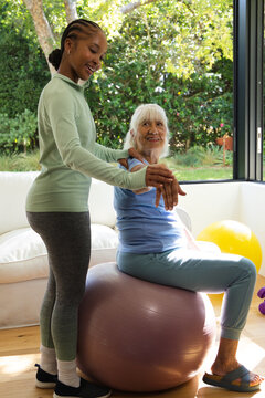 Senior woman and African American female trainer stretching on purple ball at home in activewear