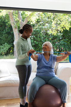 African American aide guiding senior sitting on purple ball at home holding blue resistance band