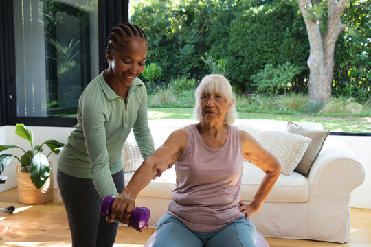 Senior woman on Swiss ball holding purple weight, African American trainer in green helping at home