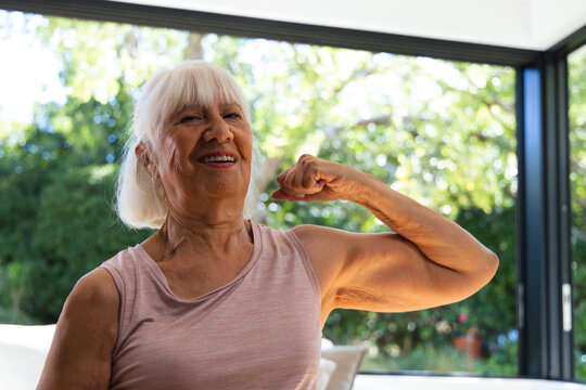 Senior female in sleeveless pink top smiling, flexing right arm while sitting on sofa by window