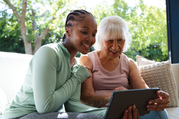 Fototapeta premium African American woman wearing green top leaning into senior woman holding tablet on sofa at home