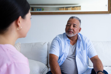 Fototapeta premium Senior African American man, Asian woman chatting on white couch, man wearing blue shirt and watch